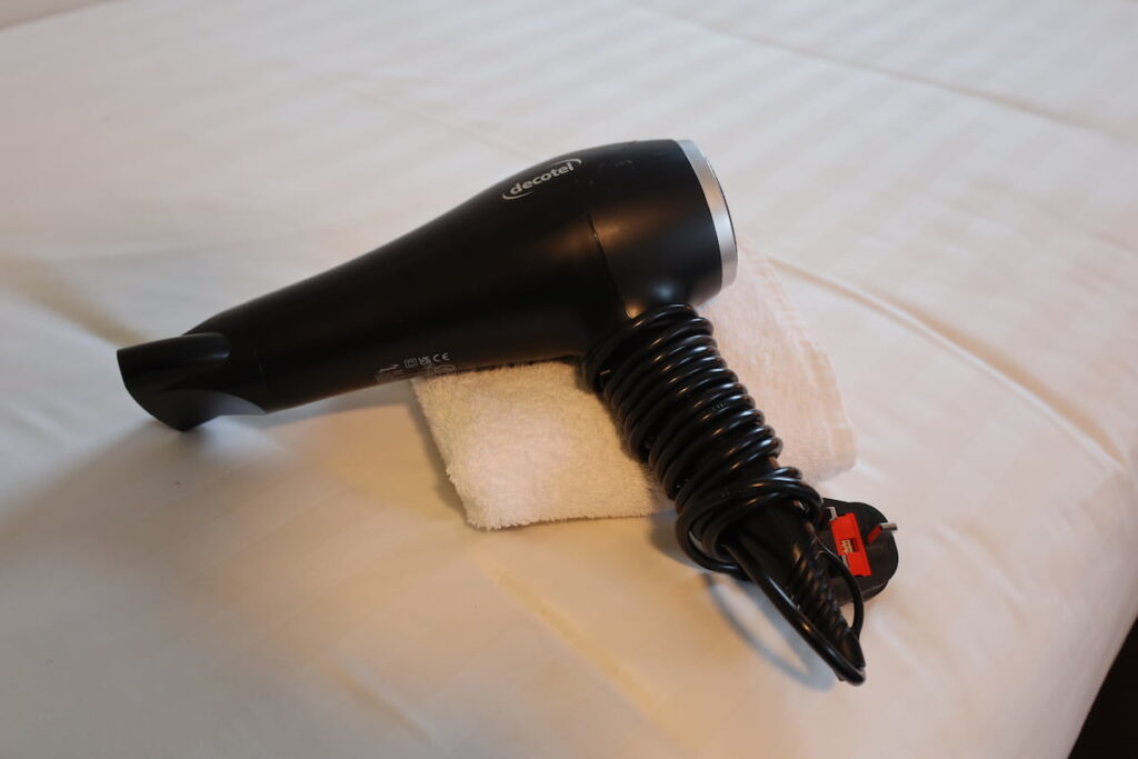 A black hair dryer with a coiled cord rests on a folded white towel on a bed with white striped sheets in a cozy triple room Dublin visitors will love.