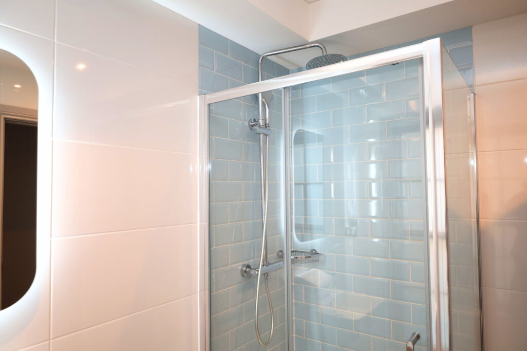 Modern shower in a triple room Dublin setting, featuring a glass door, light blue subway tiles, overhead rainfall showerhead, and a wall-mounted handheld showerhead visible.