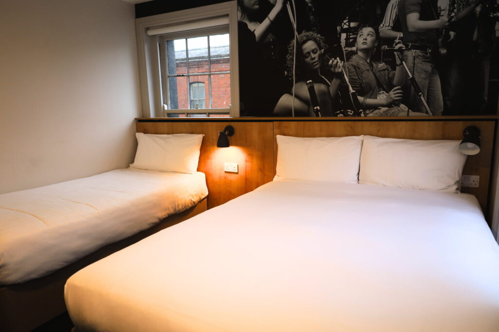 A stylish triple room Dublin features one double bed and one single bed with crisp white bedding, a wooden headboard, wall-mounted lights, and a striking black-and-white photo above the beds.