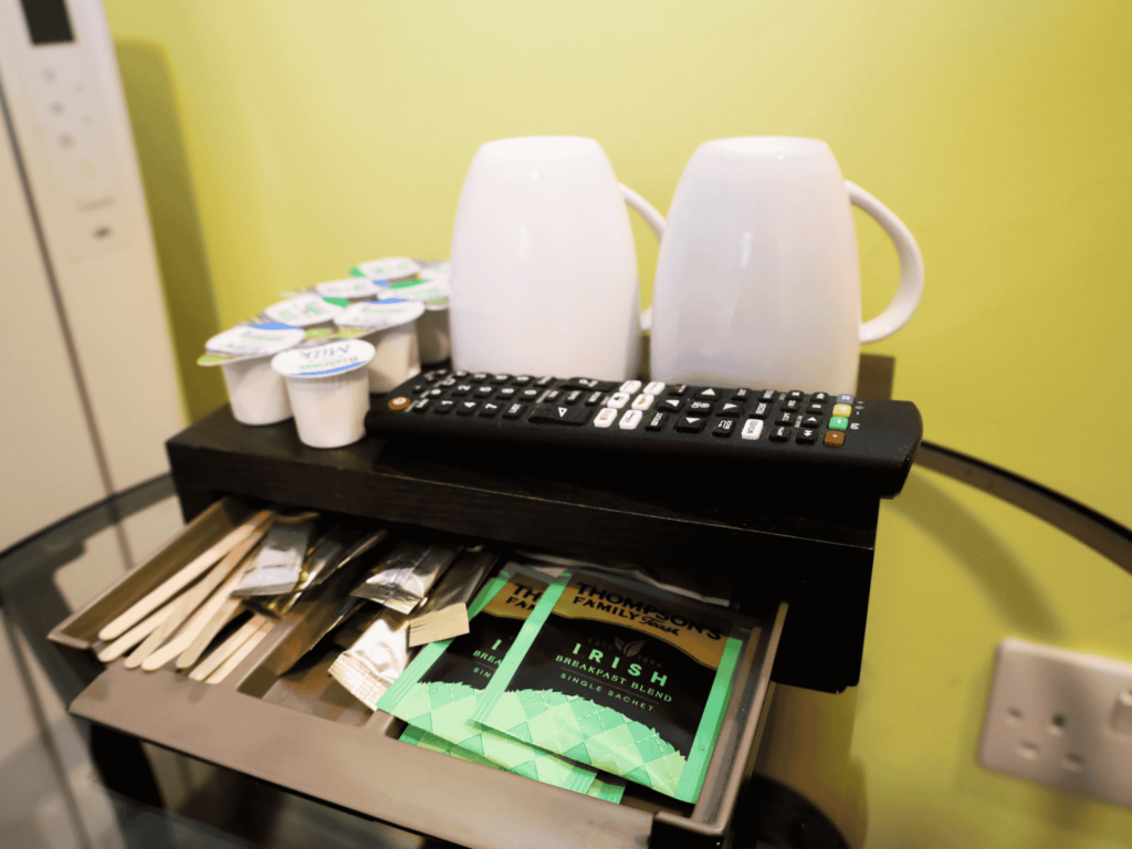 Two white mugs, a TV remote, coffee creamers, and tea bags are arranged on a tray with an open drawer containing stirrers and sweeteners on a glass table.