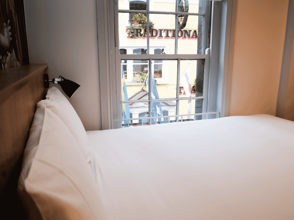 A neatly made bed with white linens next to a window, through which a building with the word “TRADITIONA” on its sign is visible.