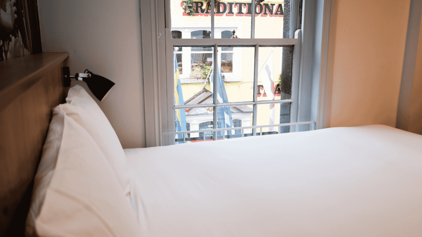 A neatly made bed with white linens next to a window, through which a building with the word &ldquo;TRADITIONA&rdquo; on its sign is visible.