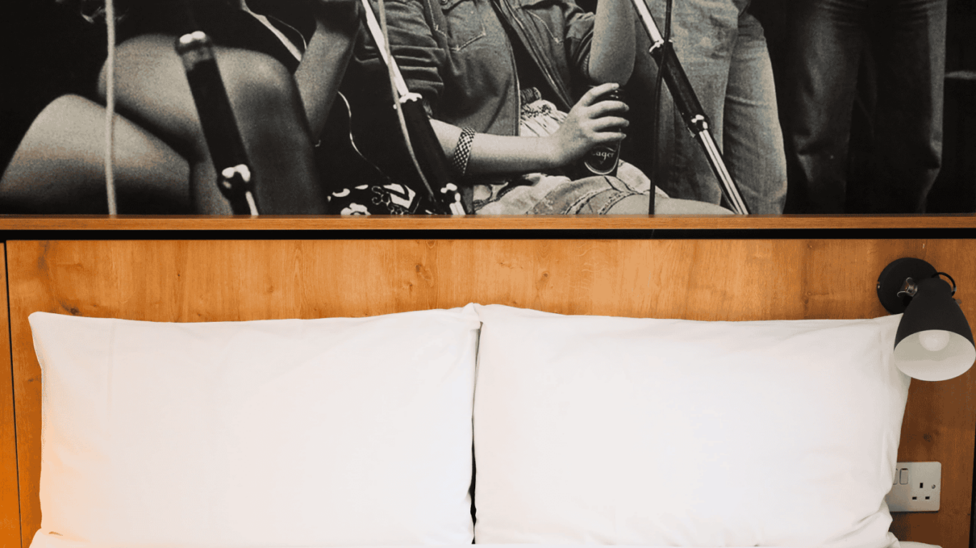 A neatly made bed with two white pillows and a wooden headboard; a black and white photo of musicians with microphones hangs above the bed.
