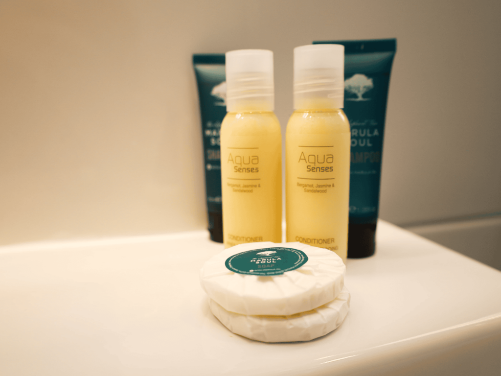 Two bottles of Aqua Senses conditioner and two round wrapped soaps are placed on a white bathroom surface, with two teal shampoo tubes in the background.