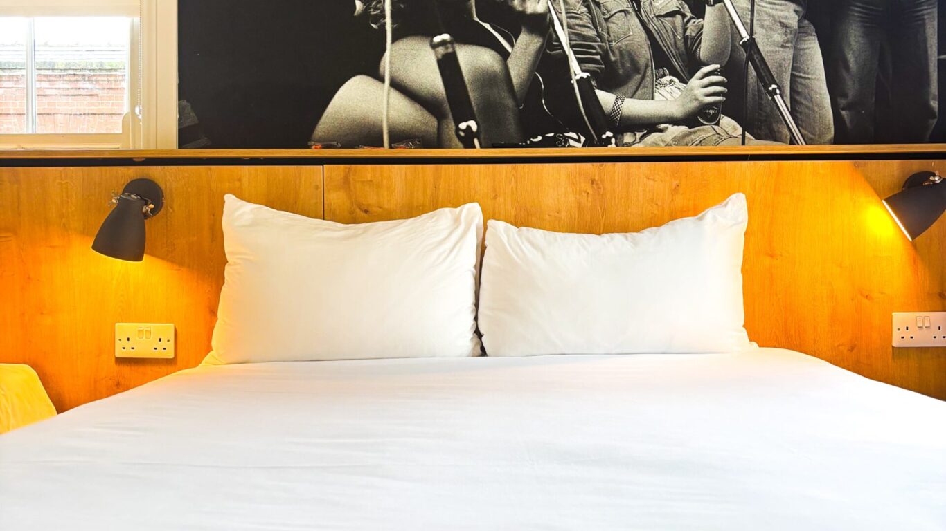 A neatly made bed with white sheets and pillows, set against a wooden headboard, with a large black-and-white photo of musicians on the wall above.