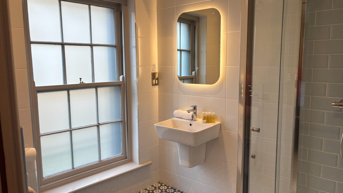 Modern bathroom with frosted window, wall-mounted sink, illuminated rectangular mirror, glass shower enclosure, and patterned floor tiles. Toiletries are placed on the sink.