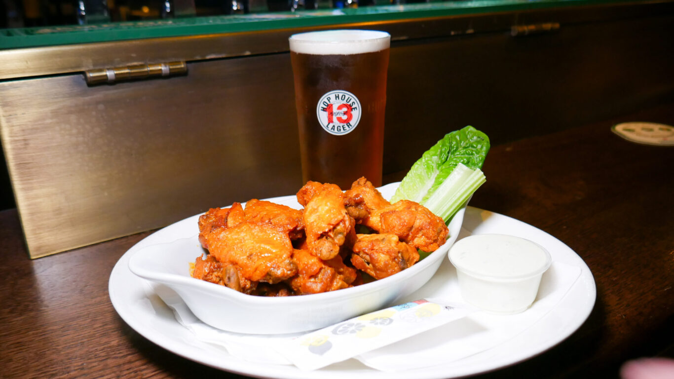 A plate of buffalo chicken wings with celery, ranch dip, and a pint of beer on a wooden table at a bar.