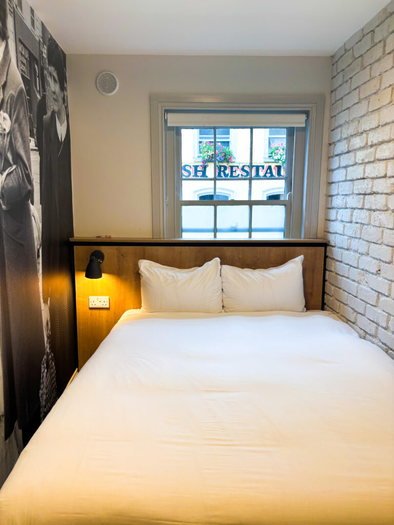 A small, neatly made bed with white linens is positioned under a window in a room with a brick accent wall and a black-and-white mural on the side.