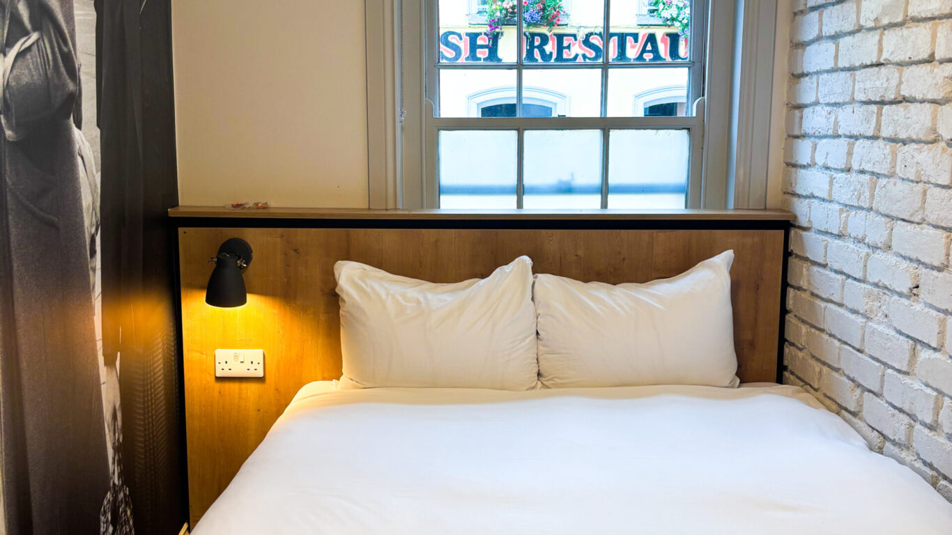 A small, neatly made bed with white linens is positioned under a window in a room with a brick accent wall and a black-and-white mural on the side.