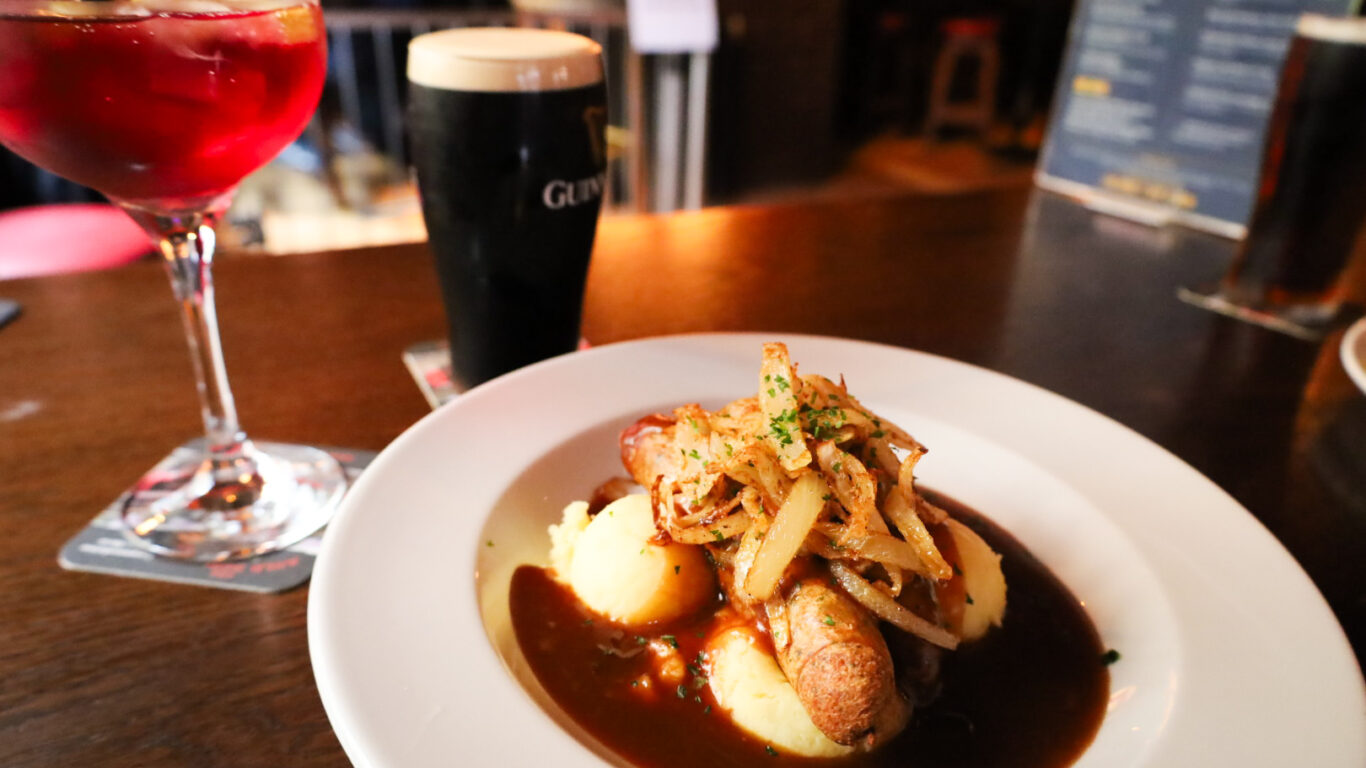 A plate of sausages with mashed potatoes and gravy is on a wooden table, accompanied by a glass of red drink and a pint of Guinness.