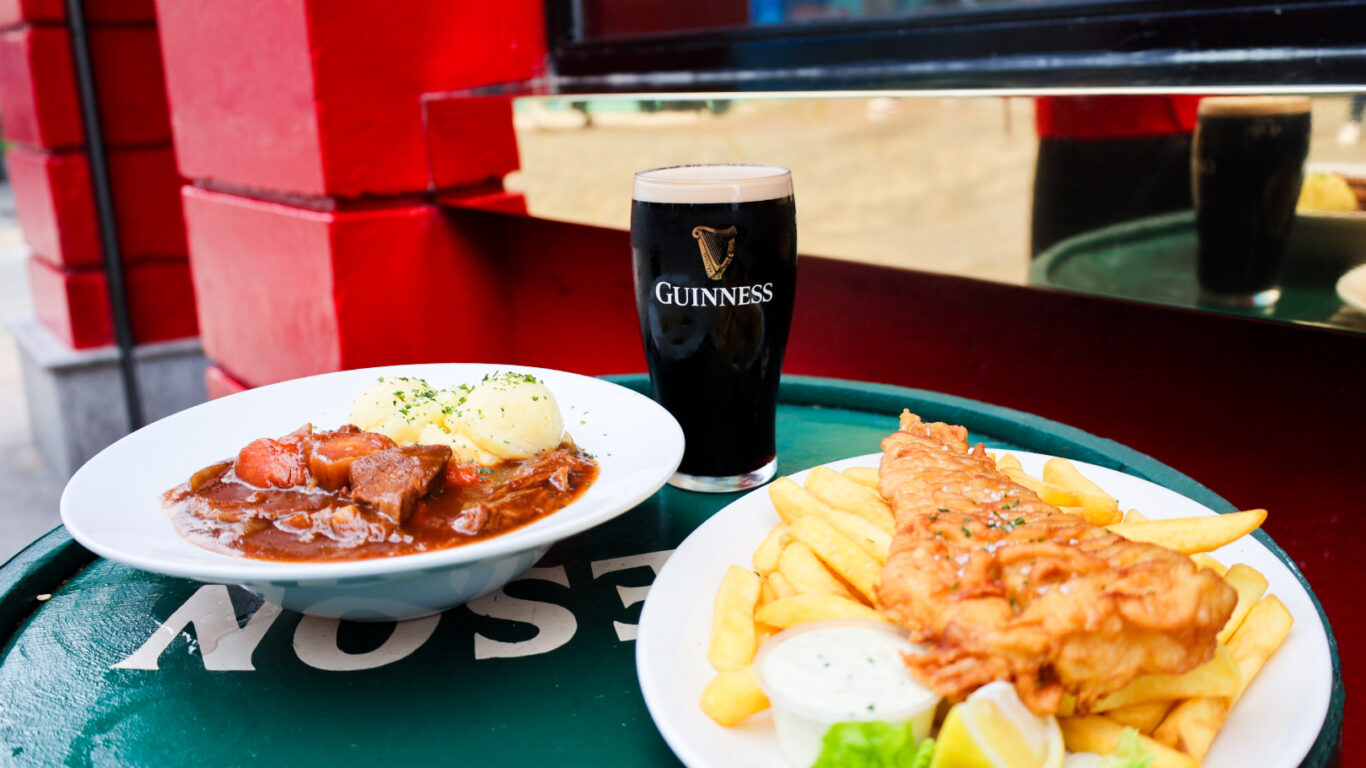A plate of fish and chips, a bowl of beef stew with mashed potatoes, and a pint of Guinness are set on a green outdoor table.