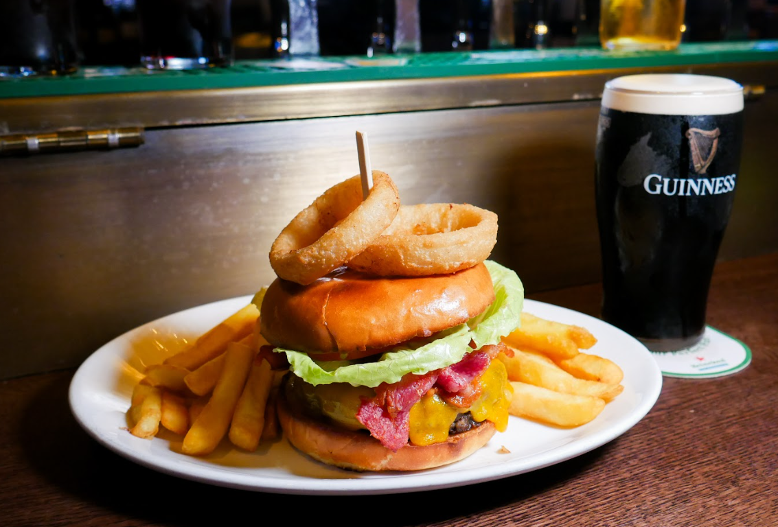 A cheeseburger with bacon, lettuce, and onion rings on a plate with fries, next to a pint of Guinness on a bar counter.