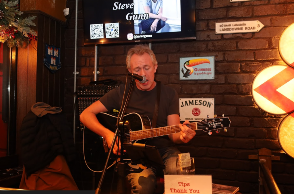 A man plays an acoustic guitar and sings into a microphone at a pub, with signs and tip jars visible around him and a screen behind displaying 