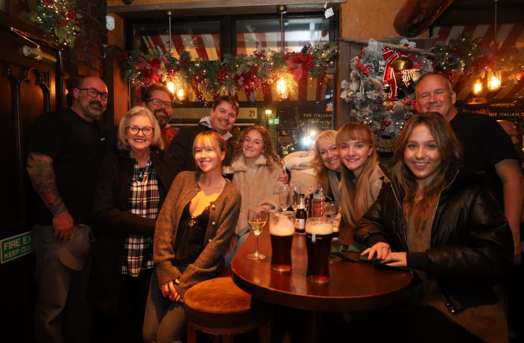 A group of people pose and smile for a photo inside a dimly lit, decorated pub with drinks on the table.