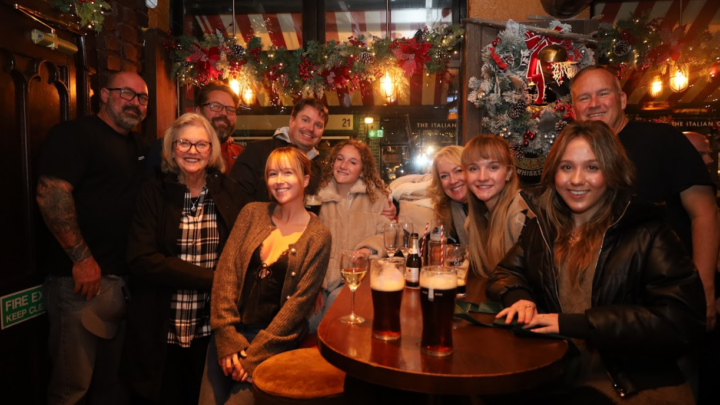 A group of people pose and smile for a photo inside a dimly lit, decorated pub with drinks on the table.