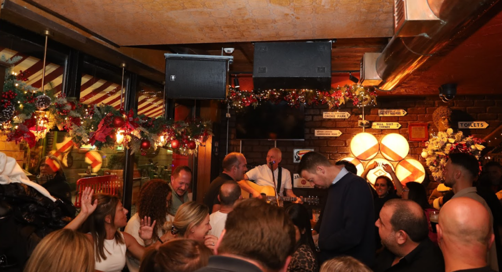 A crowd watches a musician perform on guitar in a decorated bar with Christmas ornaments and garlands.