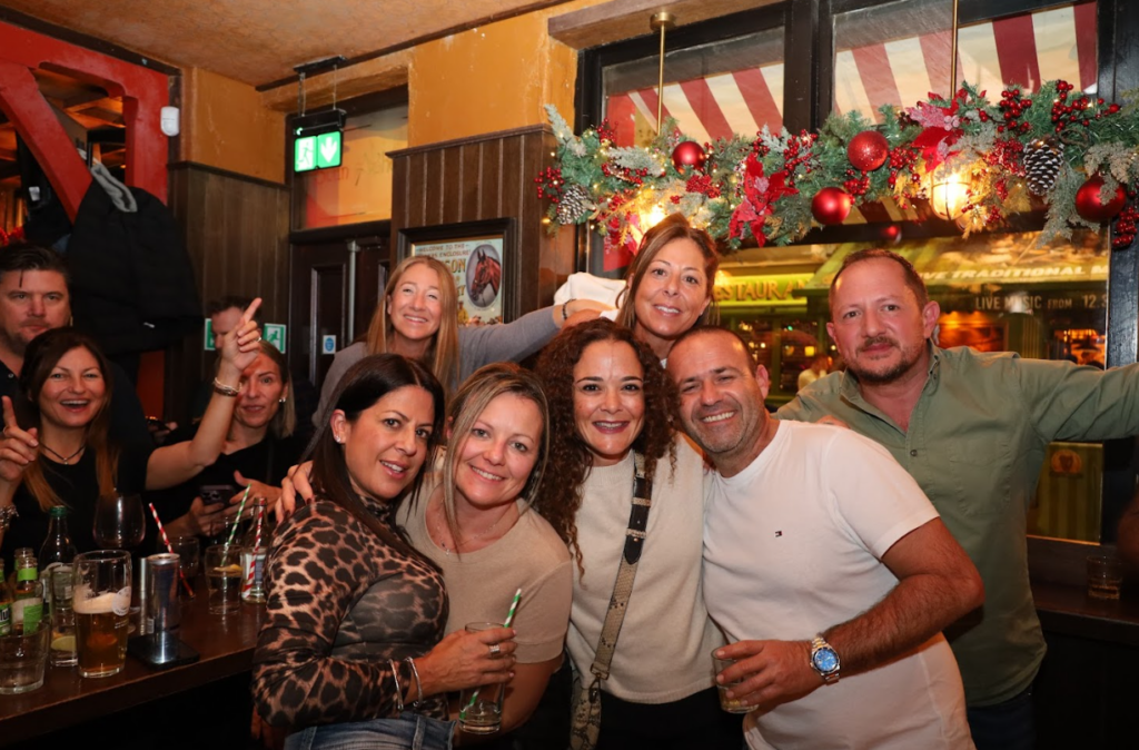A group of people posing and smiling together in a lively bar decorated with festive garland, drinks on the table in front of them.