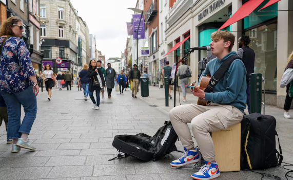 A young man plays guitar and sings into a microphone on a busy city street in Dublin&mdash;one of the Best Things to do in Dublin&mdash;while people walk by and open instrument cases lie on the ground.