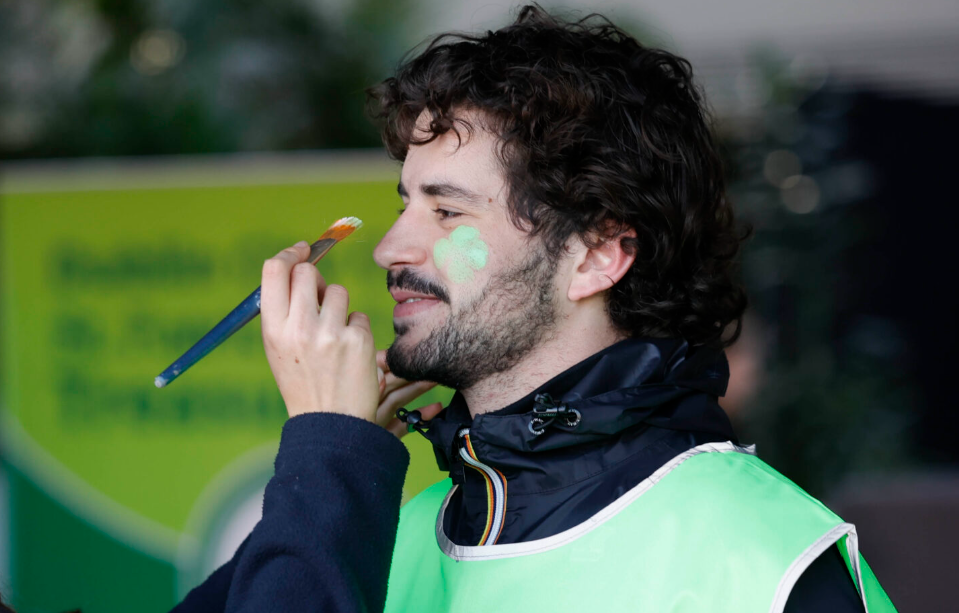 A person is having a green shamrock painted on their cheek with a brush, wearing a green vest and smiling&mdash;a fun activity often enjoyed during celebrations and one of the many festive things to do in Dublin.