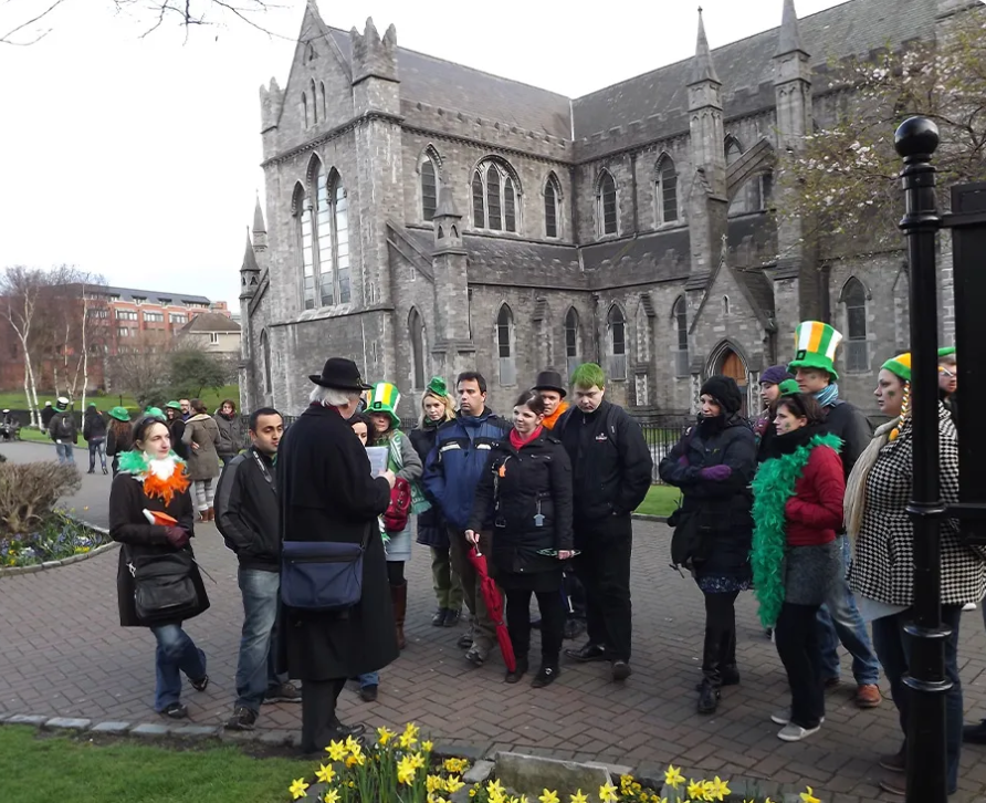 A tour group gathers outside a historic stone church, with several people wearing green hats and accessories for St. Patrick&rsquo;s Day&mdash;one of the festive things to do in Dublin.