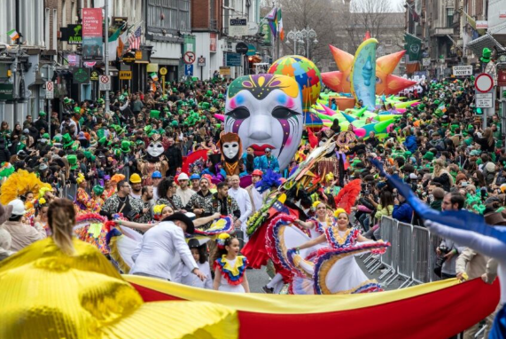 A crowded street parade in Dublin features colorful costumes, large masks, and vibrant floats, with spectators lining both sides&mdash;one of the must-see things to do in Dublin.