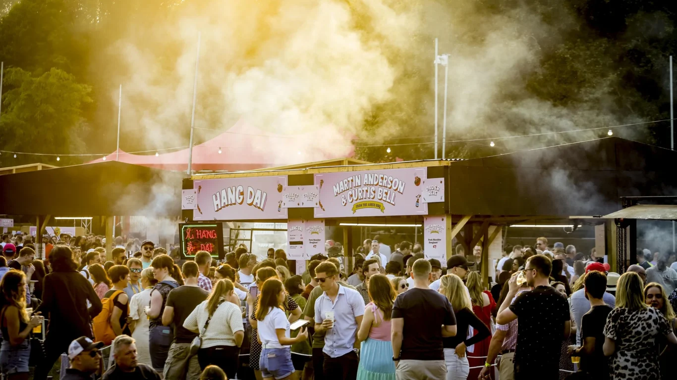 Vibrant outdoor festival scene with food stalls, lively crowd, bright signage, and warm evening glow. Ideal for social events.