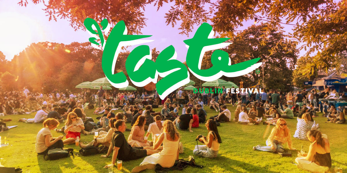 Guests enjoy a sunny day at a vibrant festival in Dublin, sitting on grass surrounded by trees, with atmosphere of joy.